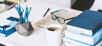 Stack of blue books on a white desk, with a coffee cup, glasses, and other office supplies